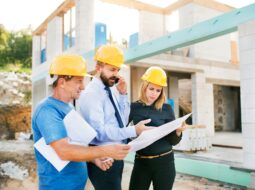 Construction professionals reviewing building plans at a residential construction site with a partially built home in the background.