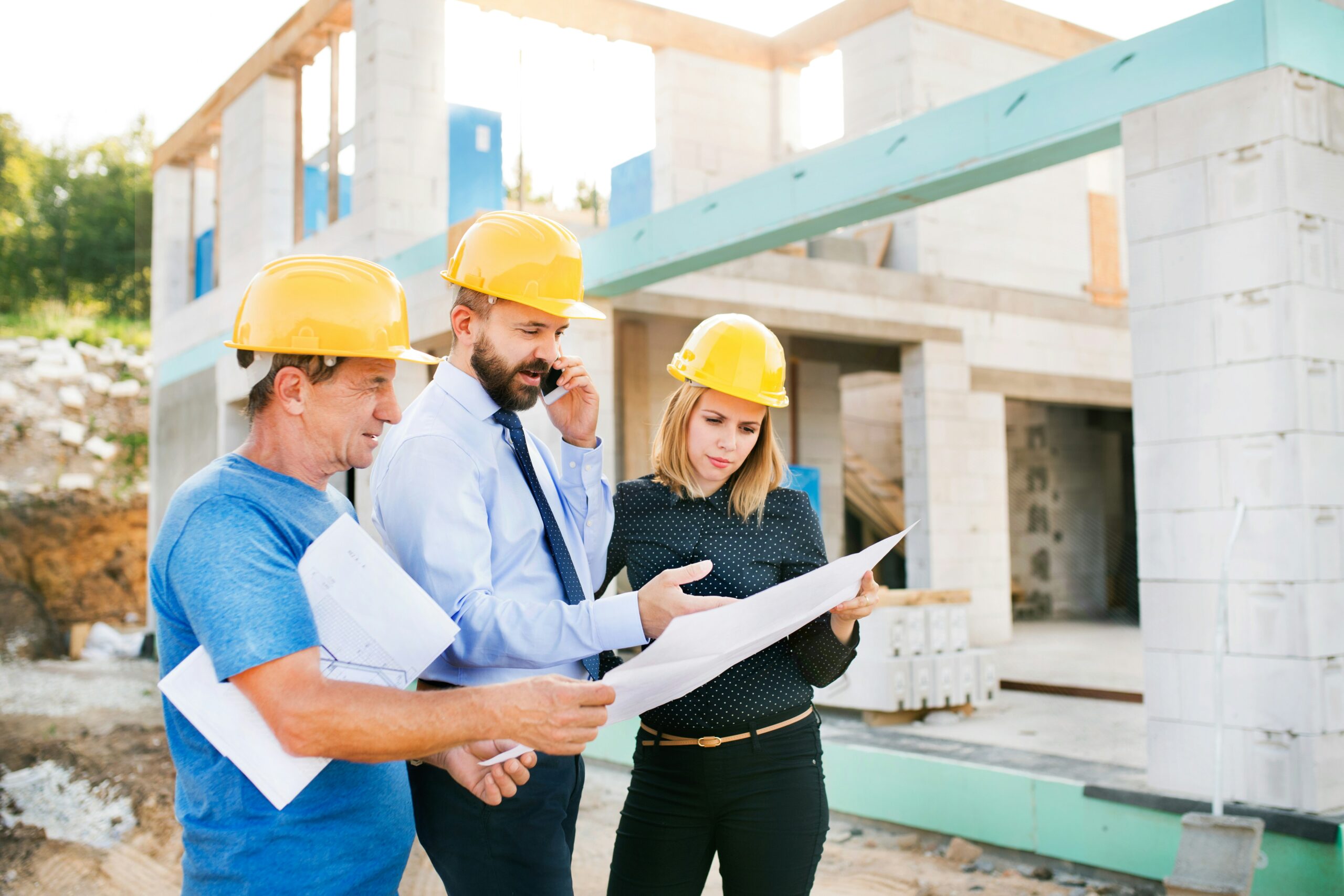 Construction professionals reviewing building plans at a residential construction site with a partially built home in the background.